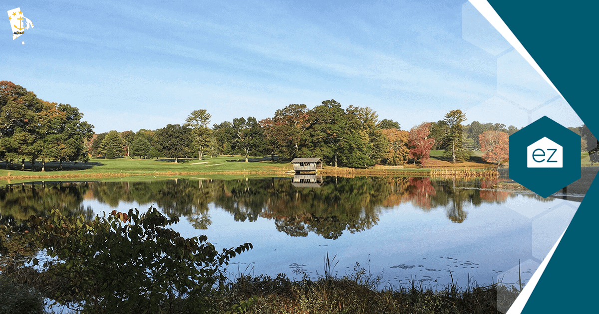 Calm Rhode Island lake reflecting autumn foliage and a small wooden structure under a clear blue sky