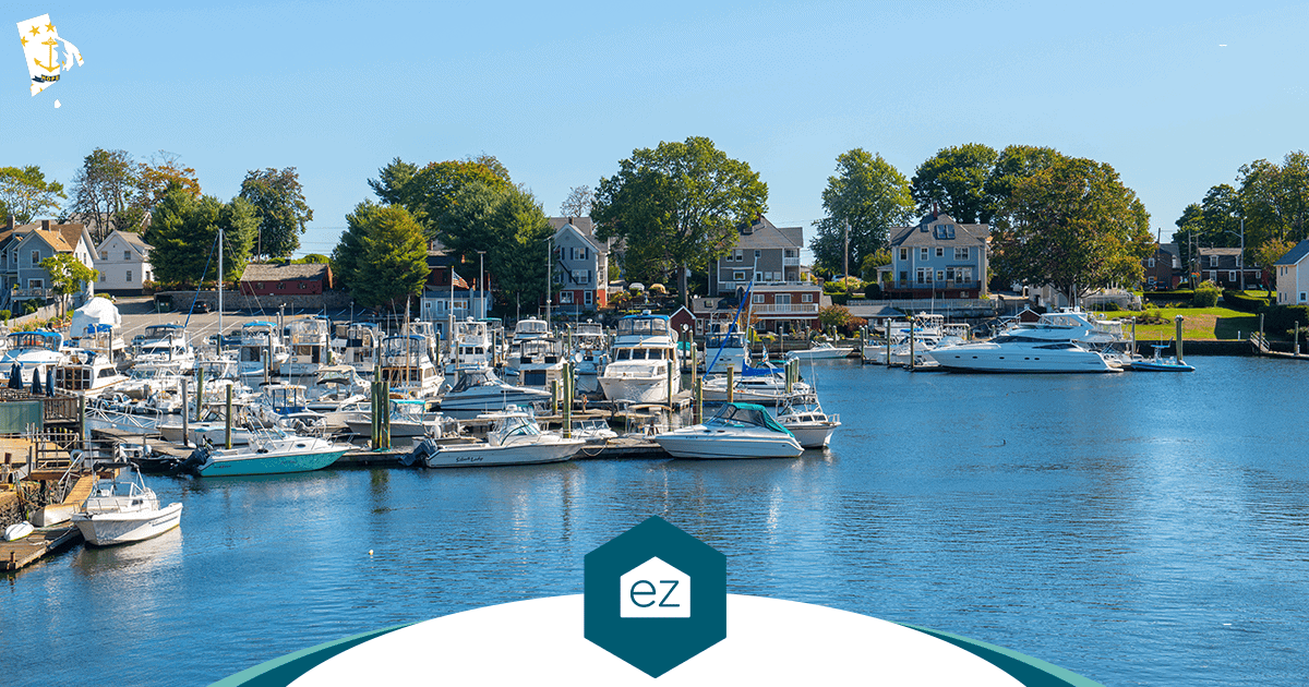 Boats docked at a small Rhode Island marina with houses and trees along the shoreline on a clear day