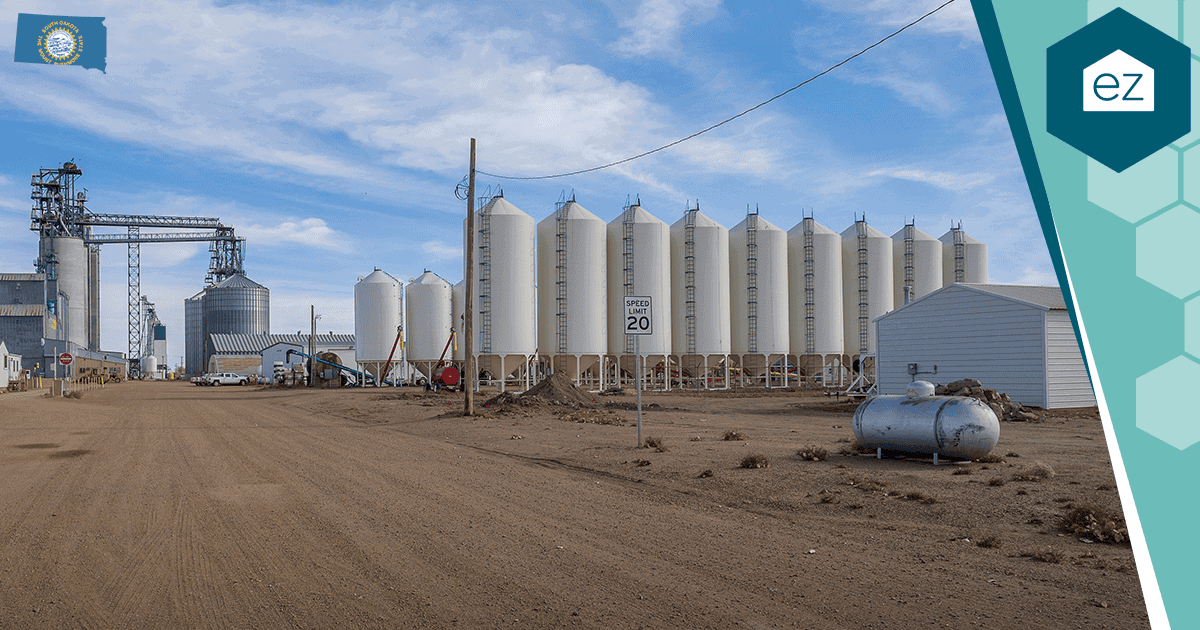 A row of large white silos stands on a dirt lot beside industrial buildings under a partly cloudy sky; a 20 mph speed limit sign is visible in the foreground, evoking the kind of landscape often sought after by those looking for unique South Dakota real estate leads.