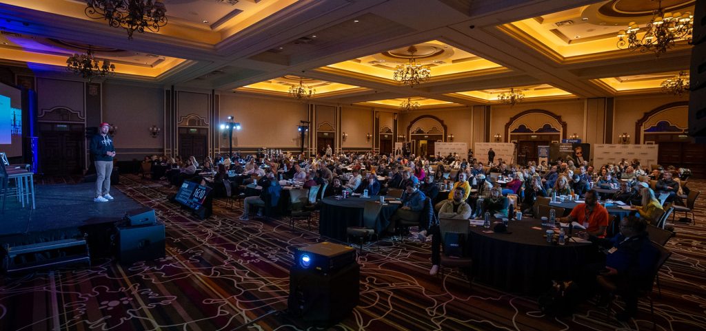 A speaker presents on stage to a large seated audience in a dimly lit conference hall with ornate ceilings and round tables, highlighting more opportunities for growth and connection.