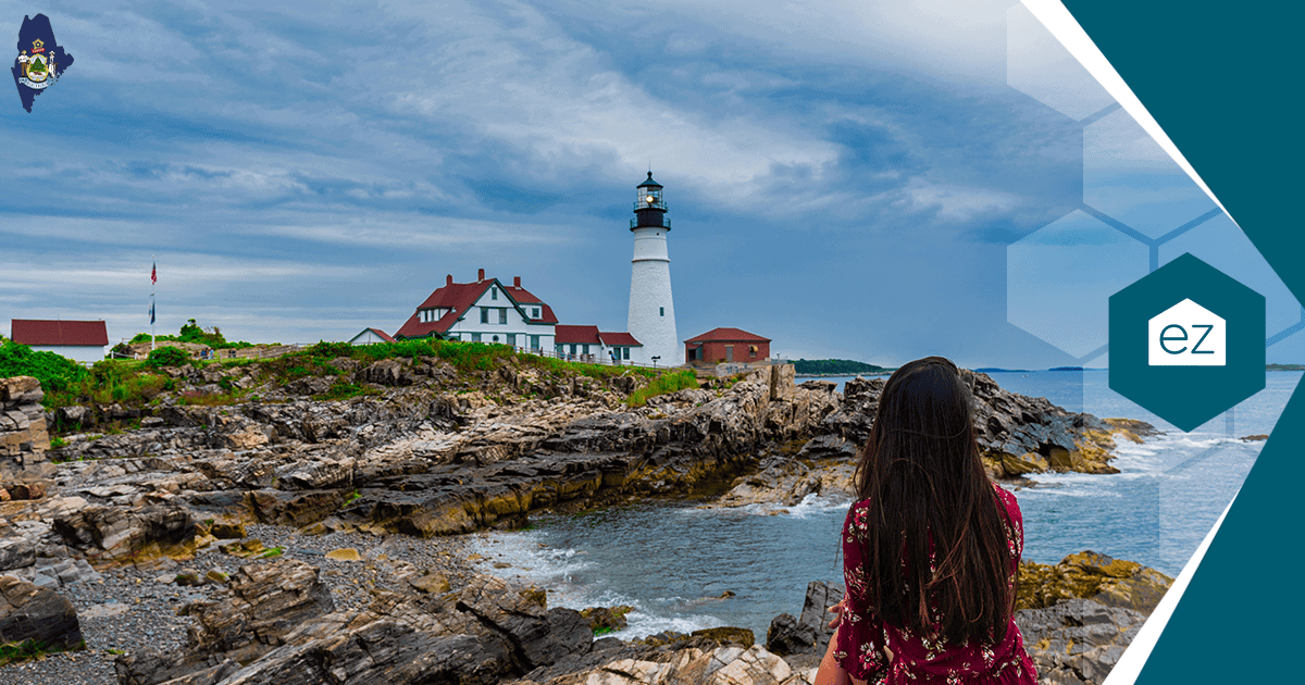 A person looks out at a white lighthouse on a rocky Maine shoreline under a cloudy sky