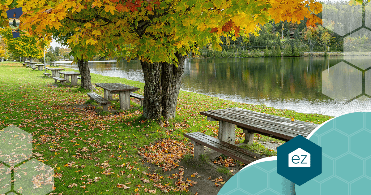 Picnic tables and benches under autumn trees beside a calm Vermont pond with forested hills behind