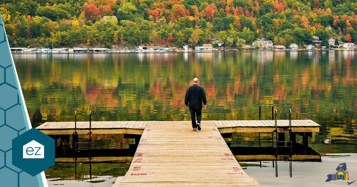 A wooden dock leading to a calm New York lake with autumn trees and lake houses on the far shore