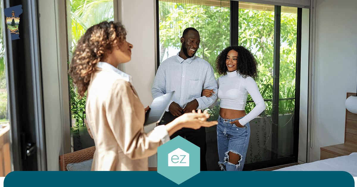 Real estate agent holding a tablet showing a bedroom to a smiling couple beside a large window with greenery outside
