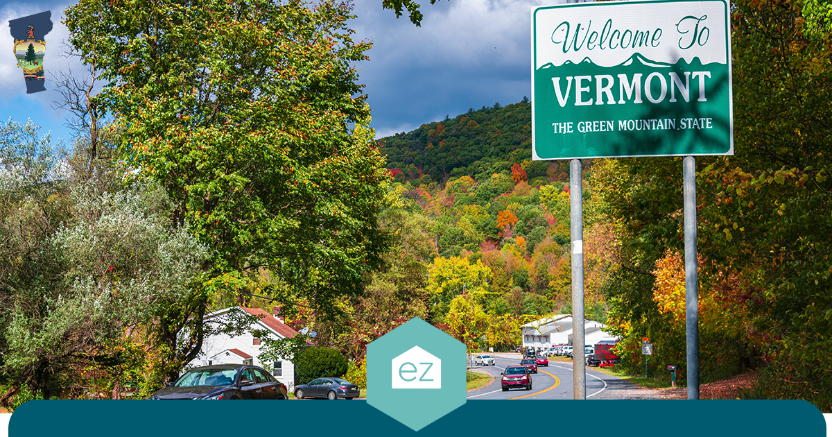 Roadside sign reading Welcome to Vermont, The Green Mountain State, beside a two-lane road with autumn foliage and houses