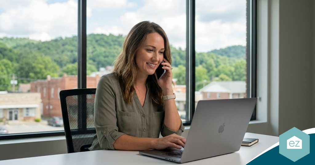 A woman smiling while talking on her smartphone at a desk with a laptop, managing her West Virginia real estate leads.
